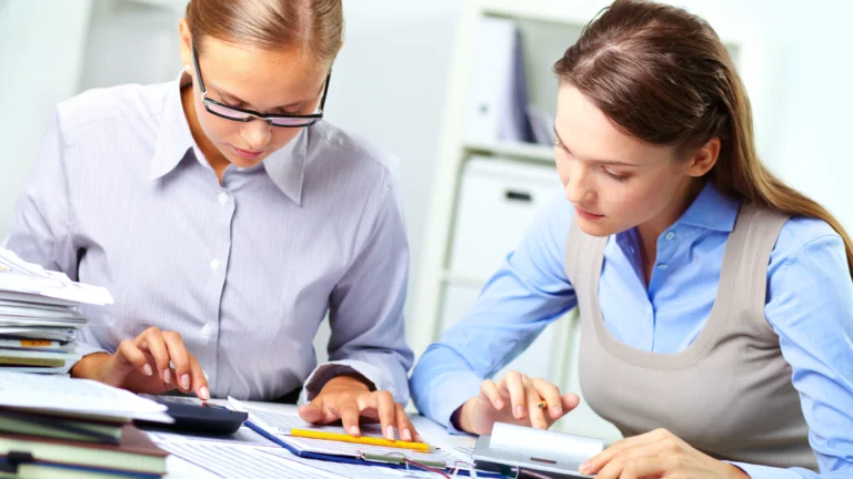 Duas mulheres, uma vereadora e sua assessora, estão concentradas em um escritório, analisando documentos financeiros detalhados. A vereadora, com óculos e vestindo uma camisa social clara, observa atentamente os cálculos em uma calculadora, enquanto sua assessora, com uma blusa azul e um colete bege, utiliza outra calculadora e examina os papéis sobre a mesa. A cena sugere um momento crucial de análise e tomada de decisão em relação às peças orçamentárias de 2025, destacando a importância da colaboração e do profissionalismo no contexto político.