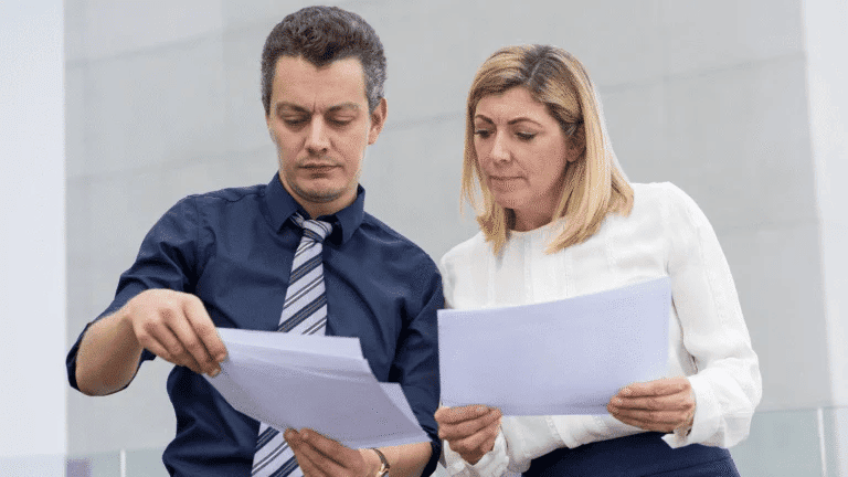 Um homem, representando um vereador, veste uma camisa escura e gravata listrada enquanto segura alguns papéis. Uma mulher, representando a prefeita, veste uma blusa clara e saia escura e analisa um dos documentos. Ambos parecem concentrados, sugerindo a análise de uma indicação parlamentar feita pelo vereador à prefeita.